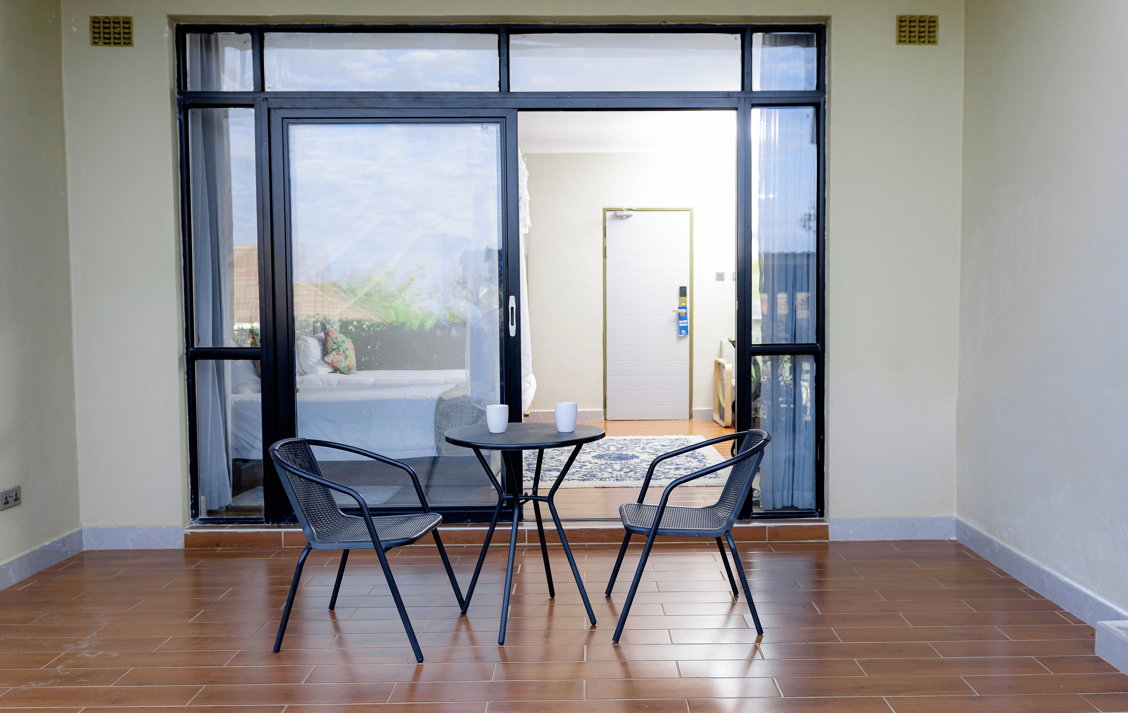 A room with a sliding glass door leading to a balcony, a small round table with two chairs, and a view of a building and greenery outside.