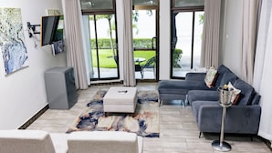 A modern living room with a grey sofa, a small ottoman, a flat-screen TV mounted on the wall, and a large window with curtains. - Sigelege Beach Resort (Senga)