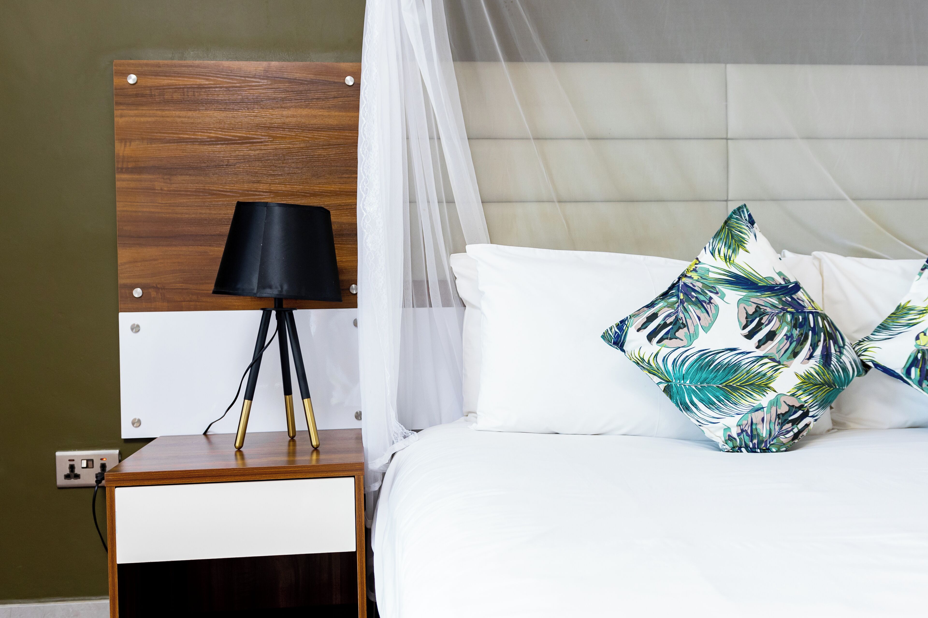 Una cama bien hecha con sábanas blancas y un cojín estampado con motivos de hojas tropicales, una mesita de noche con una lámpara negra y un cabecero de madera.