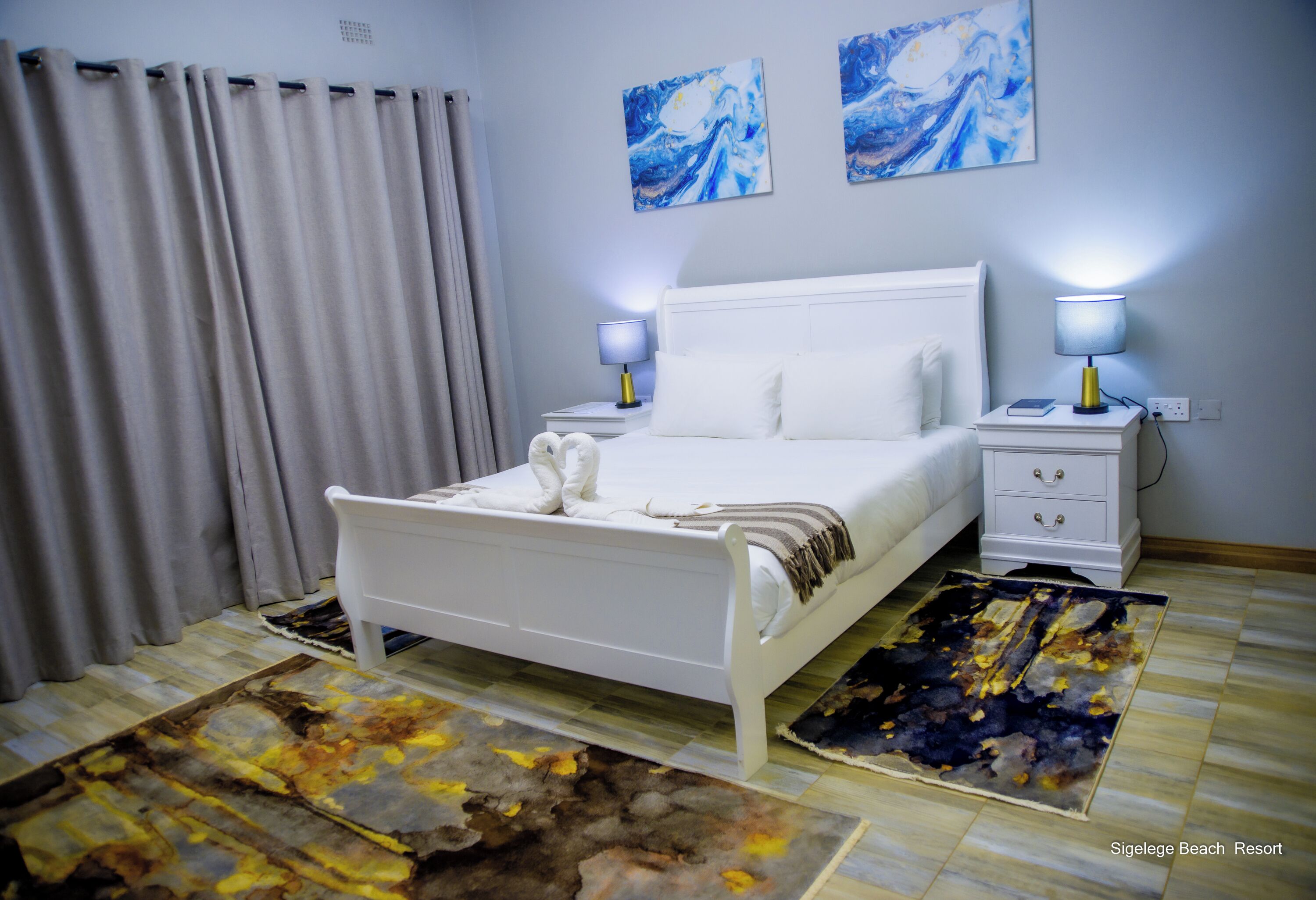 A bedroom with a white bed, two bedside tables with lamps, a large abstract rug, and two wall paintings.