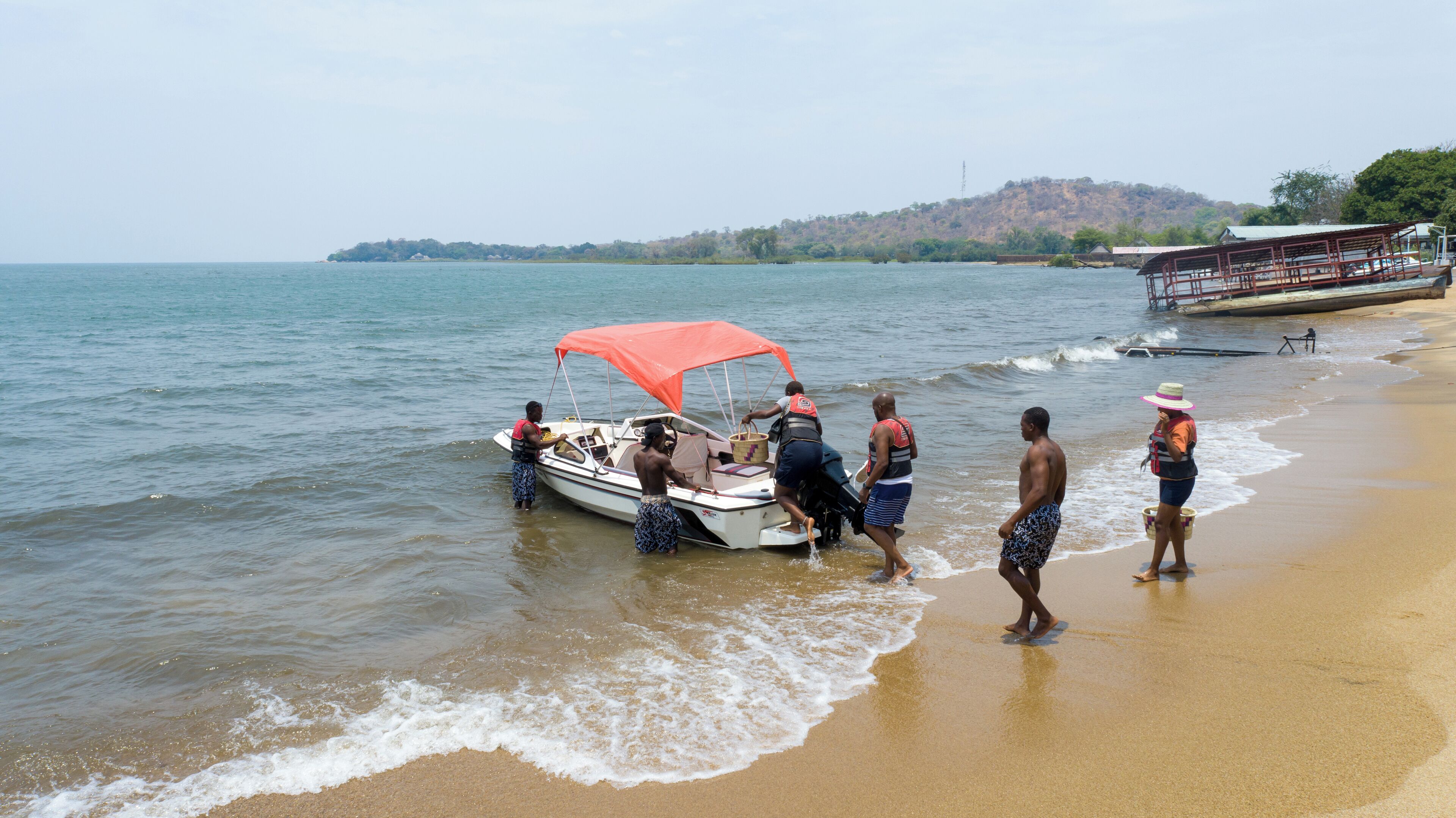 A beach scene with a boat near the shore, people walking along the beach, and a structure in the background.