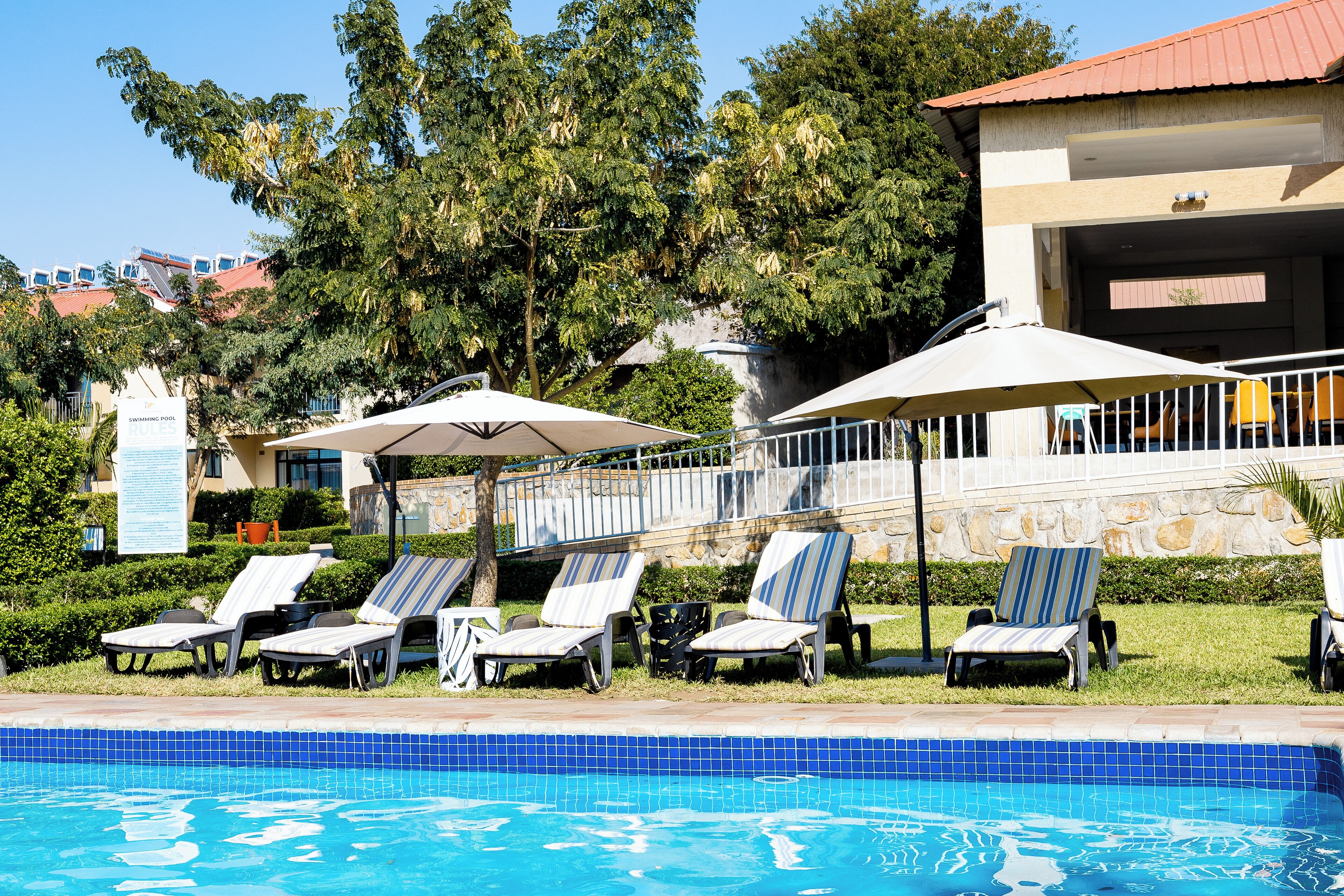 A swimming pool with lounge chairs and umbrellas, surrounded by greenery and a building with a red roof.