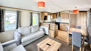 A compact RV interior with a sofa, dining table, kitchen area, and a window with curtains.