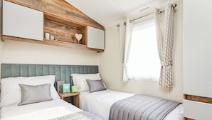 Ein Schlafzimmer mit zwei Betten, einem Holzregal mit Dekorationen, einem Fenster mit Vorhängen und einem Kopfteil.