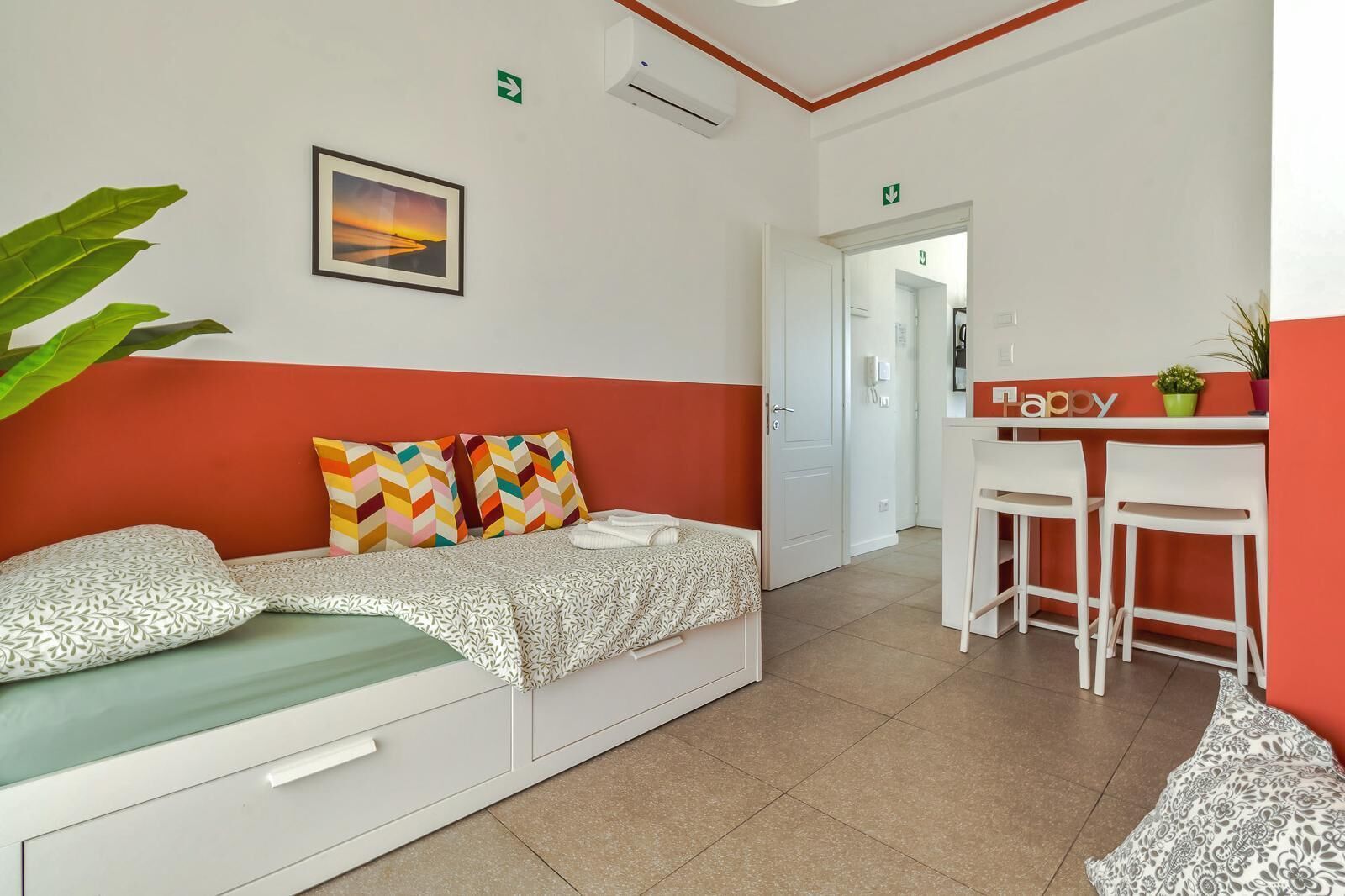 Een kamer met een bed, een bureau en een stoel. Op het bureau staat een plant en aan de muur hangt een foto.