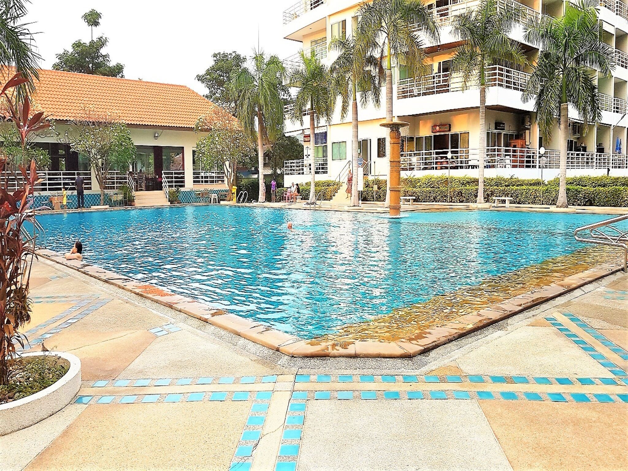 Zona de piscina con solárium de baldosas, palmeras y un edificio con balcones al fondo.