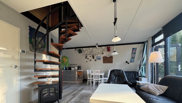 Une cuisine moderne avec un escalier en bois, un coin repas avec une table blanche et un canapé noir, et une vue sur l’extérieur grâce à de grandes fenêtres.