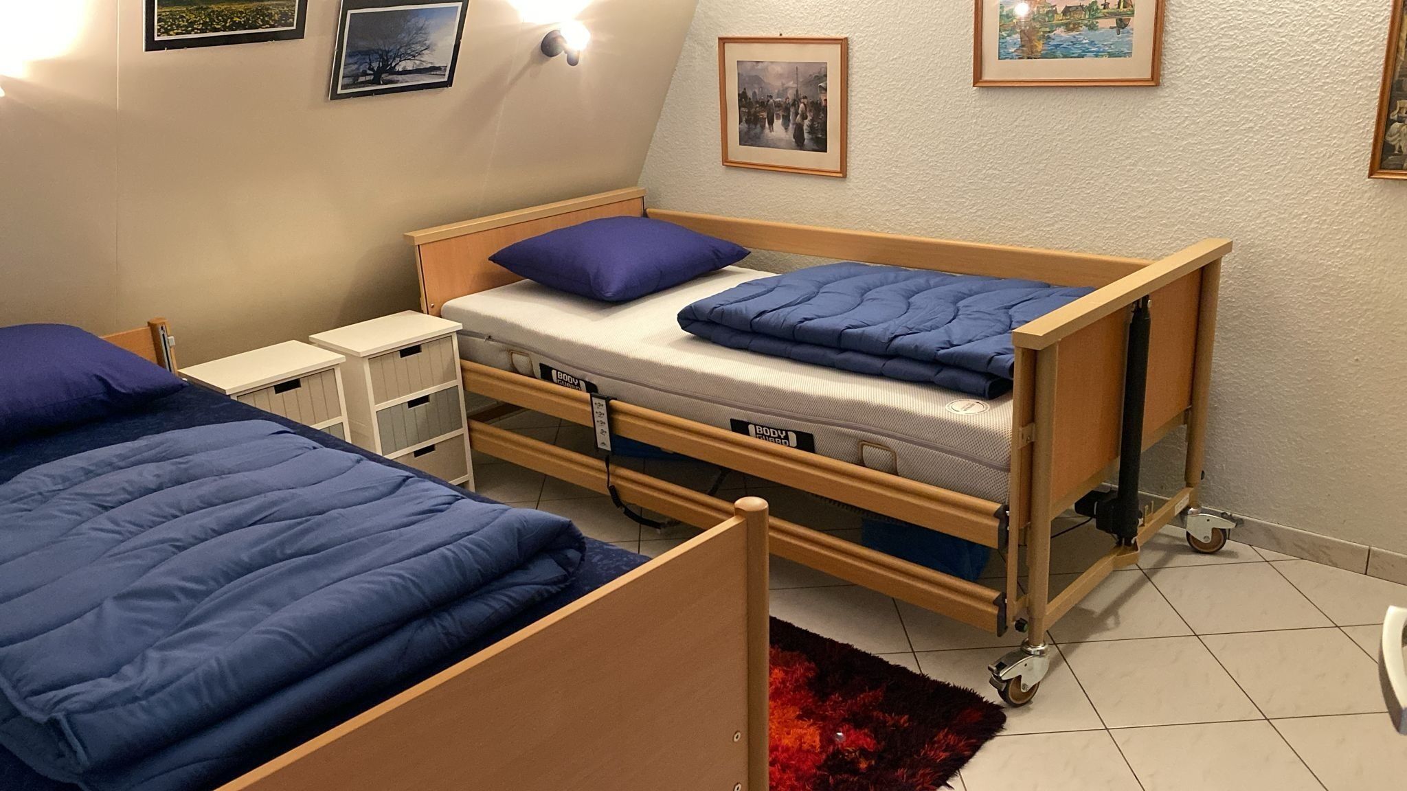 Quarto com uma cama de solteiro e uma cama de casal, ambas com roupa de cama azul. Há luminárias fixadas na parede e quadros emoldurados.