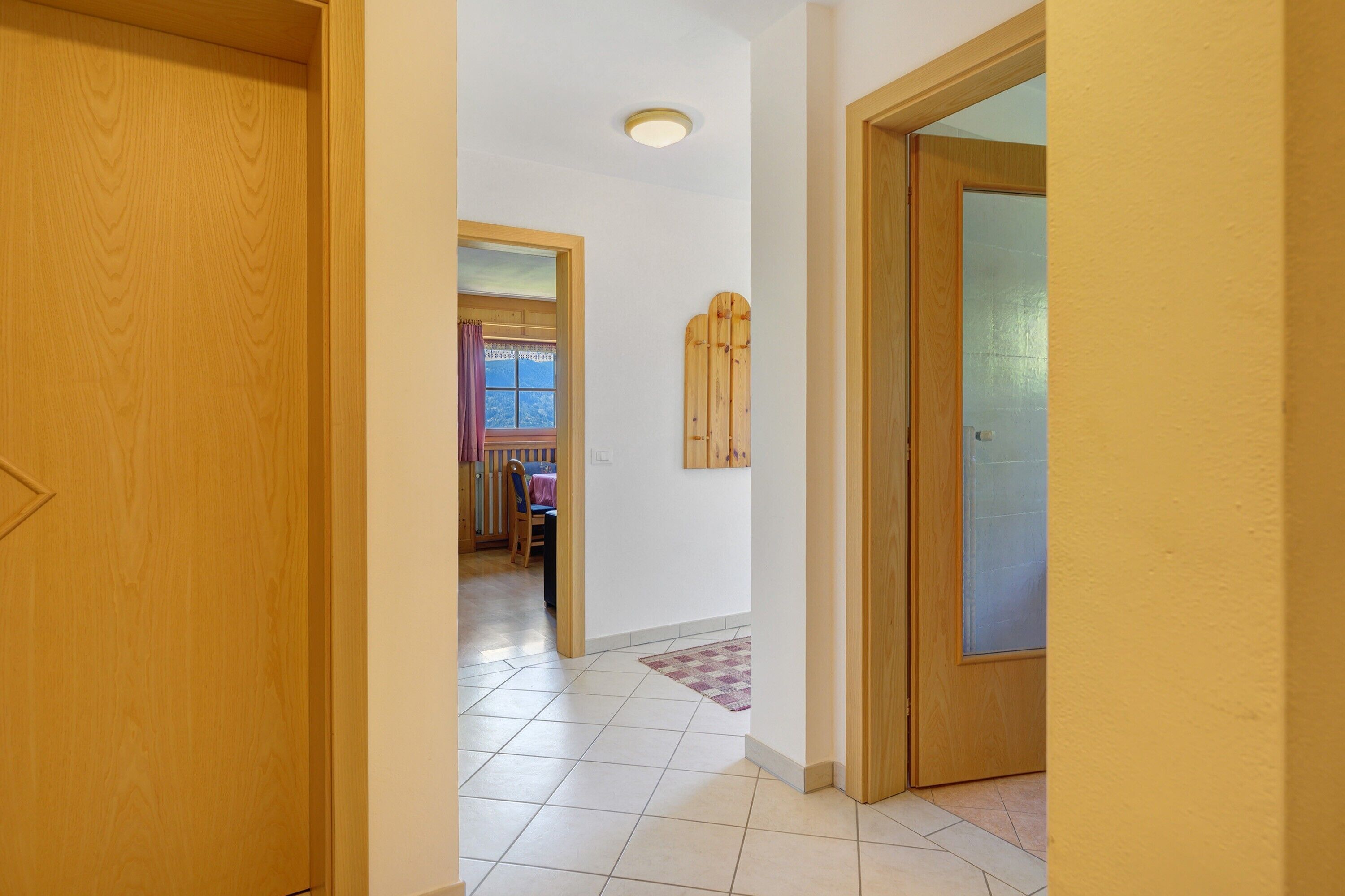 A hallway with a wooden door, a glass door, and a view into a room with a window and a wooden wall decoration.