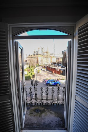 Une vue depuis une fenêtre ornée d’une rambarde en métal, donnant sur une rue bordée d’immeubles et de voitures garées.