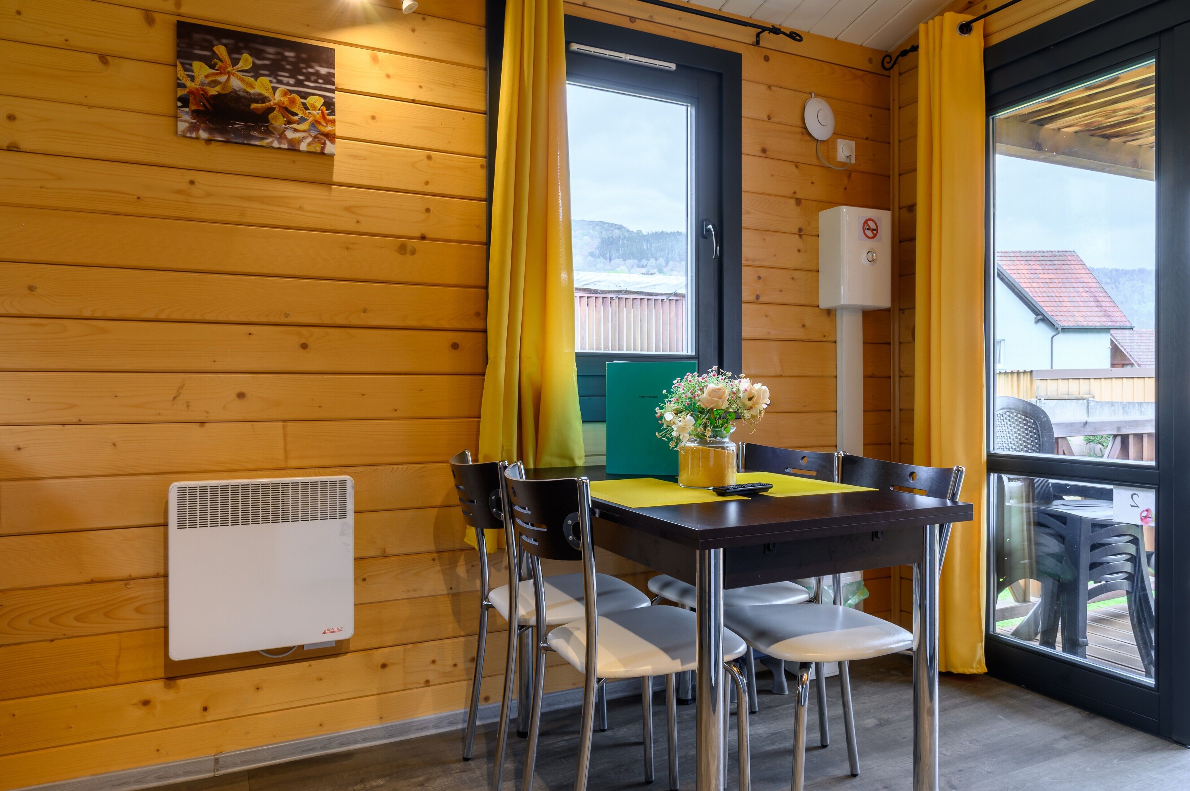Een houten interieur met een tafel, stoelen, een bloevas, een raam met gordijnen en een wandradiator.