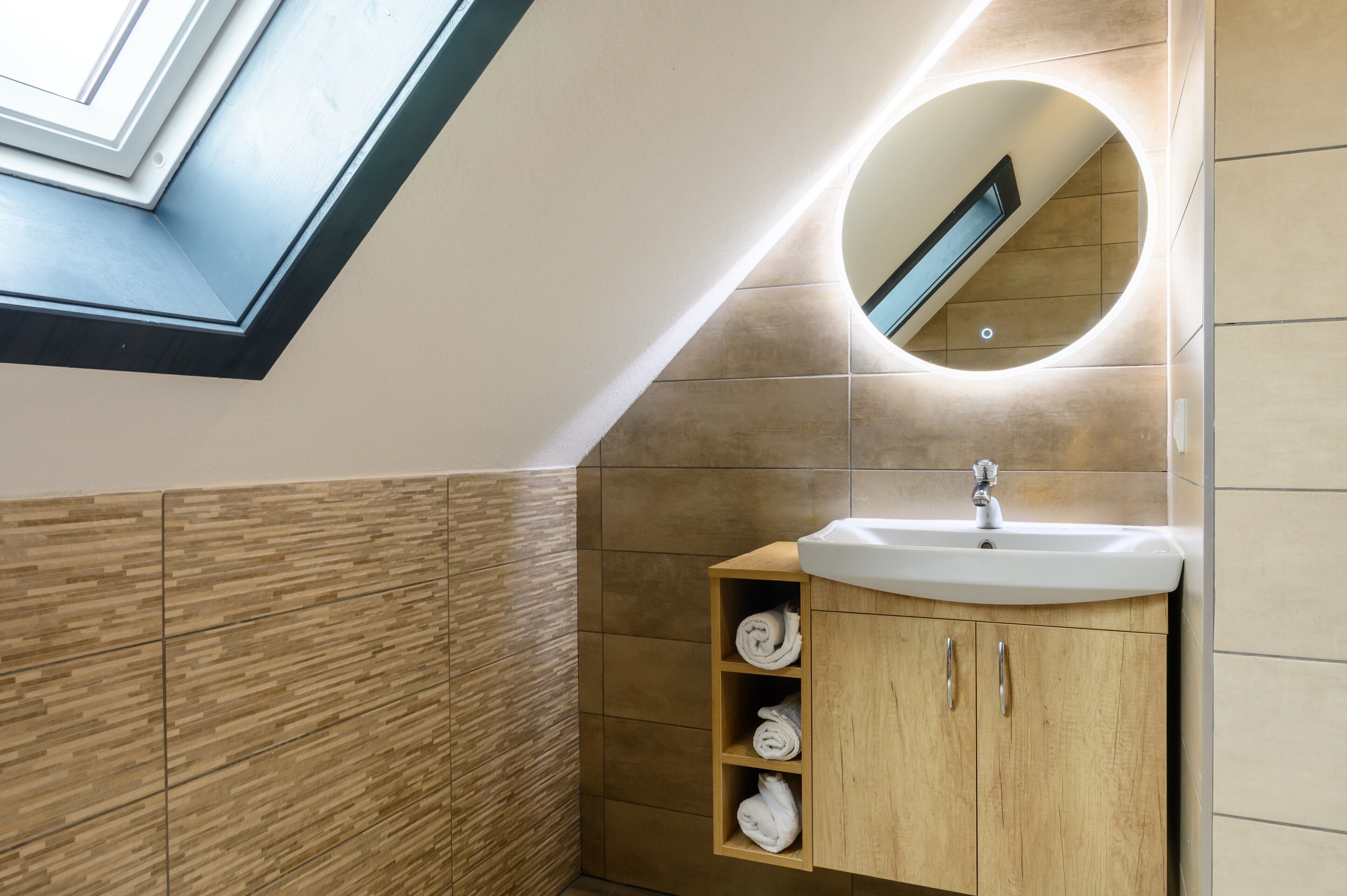 A modern bathroom with a skylight, a round mirror, a sink, and wooden cabinets.