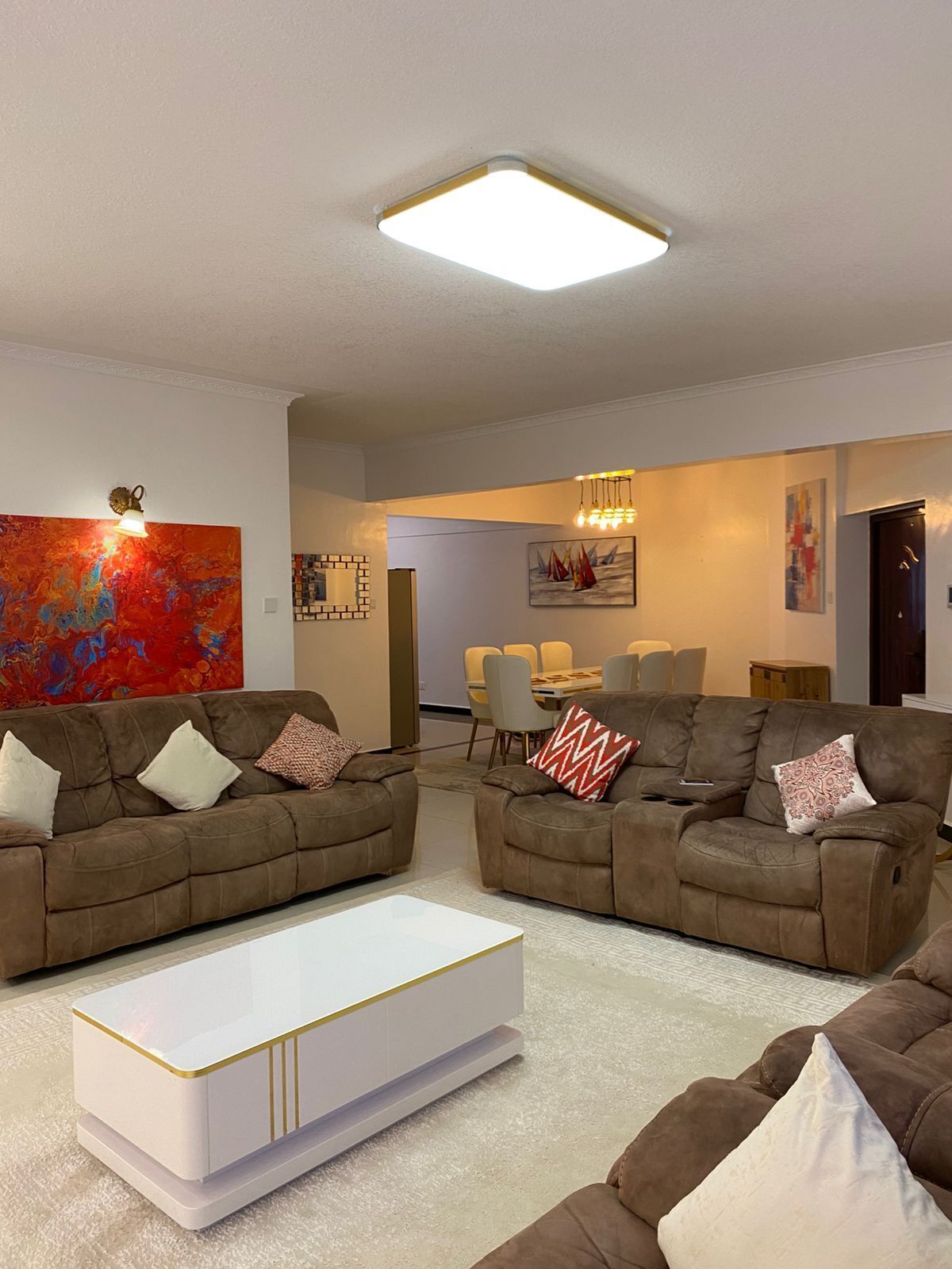 A modern living room with two brown sofas, a white coffee table, and a dining area in the background.