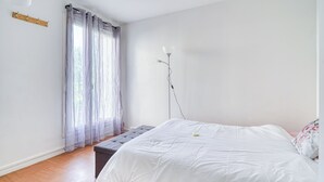 A bedroom with a bed, a bench, a bedside table, a lamp, and a window with curtains. - Cocoon rose garden (L'Haÿ-les-Roses)