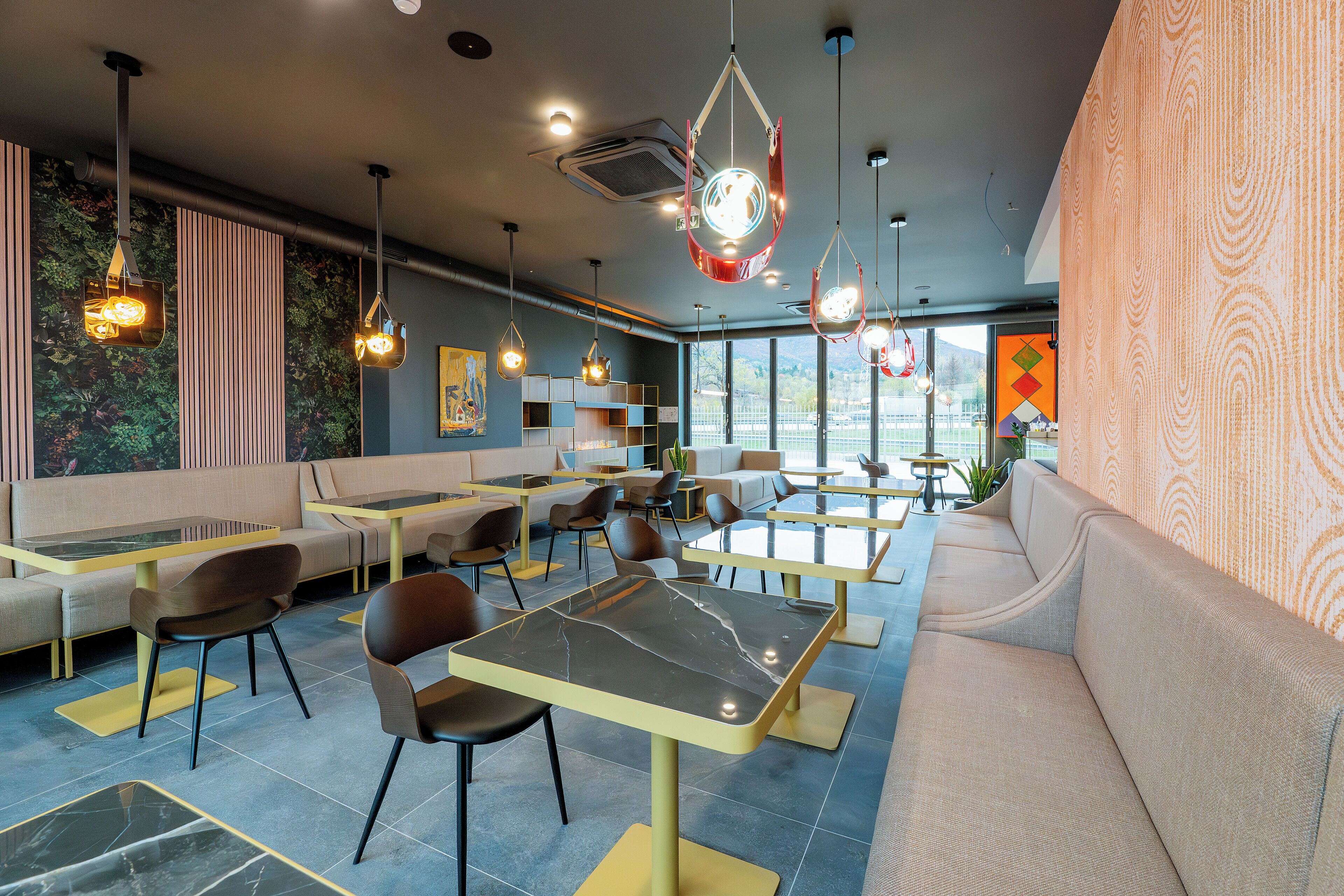 A modern restaurant interior with seating arrangements, hanging pendant lights, and a large window providing a view.