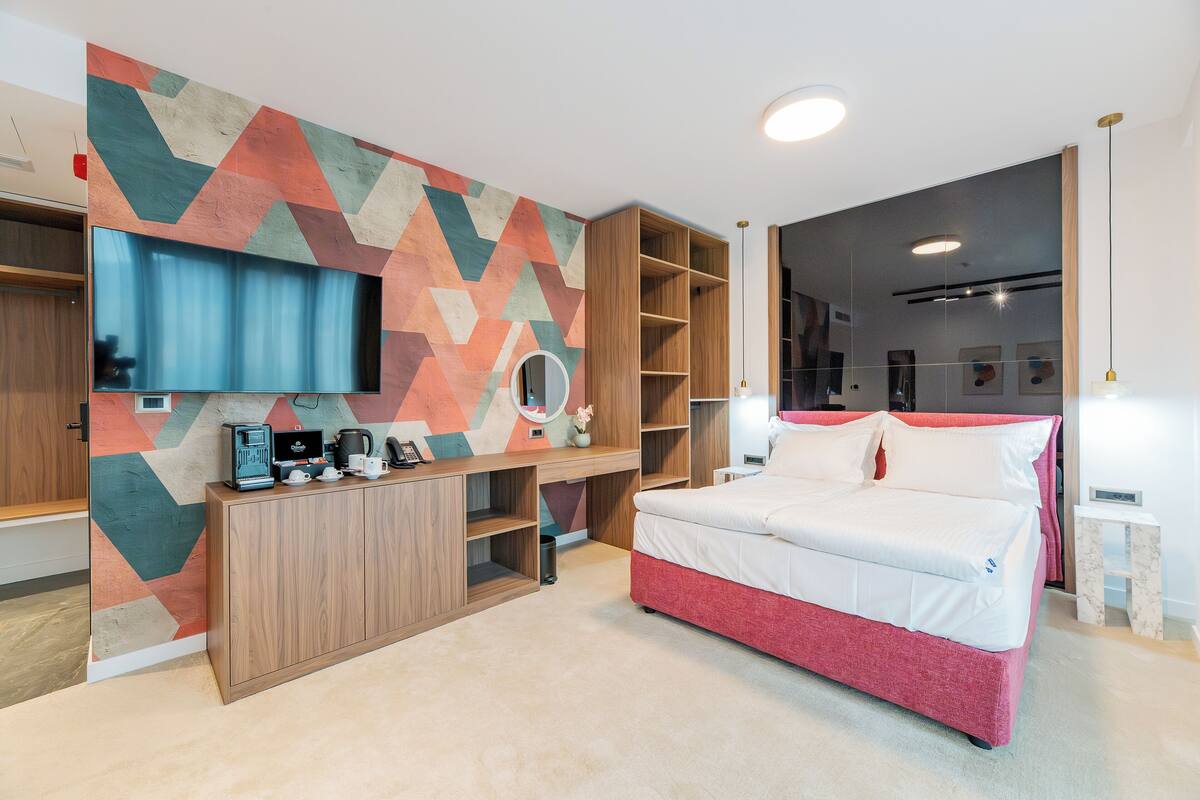 Un dormitorio moderno con cama, televisor montado en una pared con diseño geométrico, escritorio de madera con cafetera y estanterías empotradas.