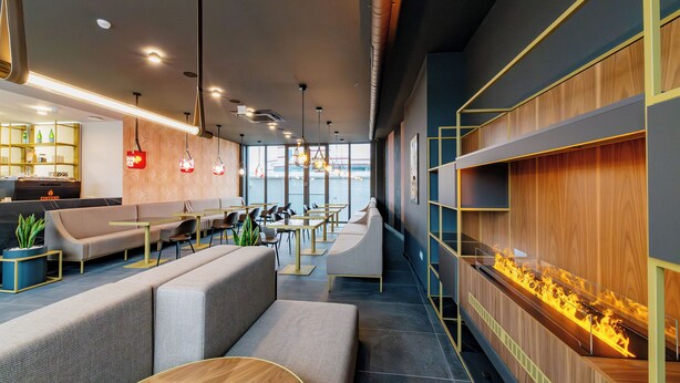 A modern interior with seating areas, a long wooden shelf with a fire feature, and a kitchen area with shelves and plants.