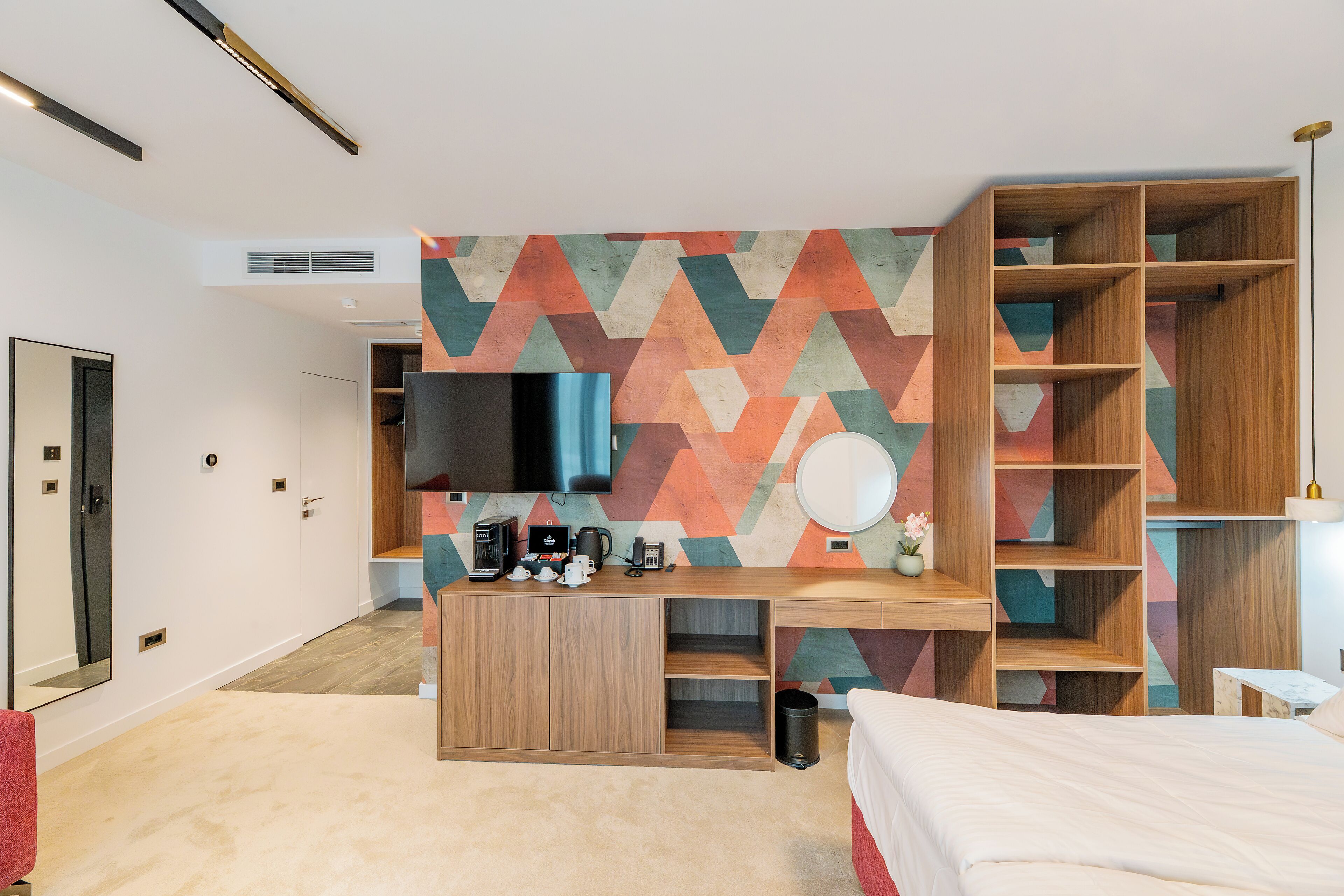 A modern hotel room with a wooden desk, a flat-screen TV, a bed, and a geometric-patterned wall.