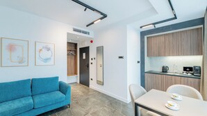 A modern hotel room with a blue sofa, a dining table with two plates, and a kitchen area with wooden cabinets.