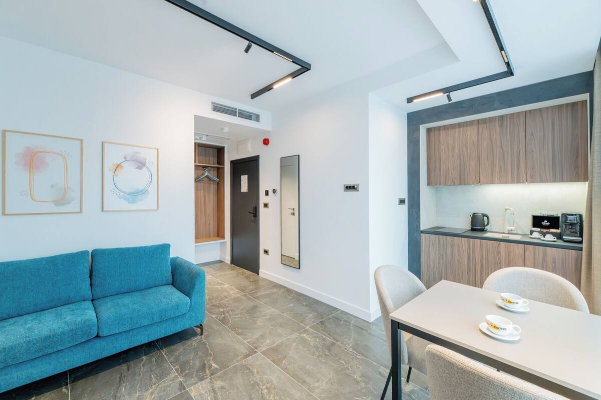 Una habitación de hotel moderna con un sofá azul, una mesa de comedor con dos platos, y una zona de cocina con armarios de madera.
