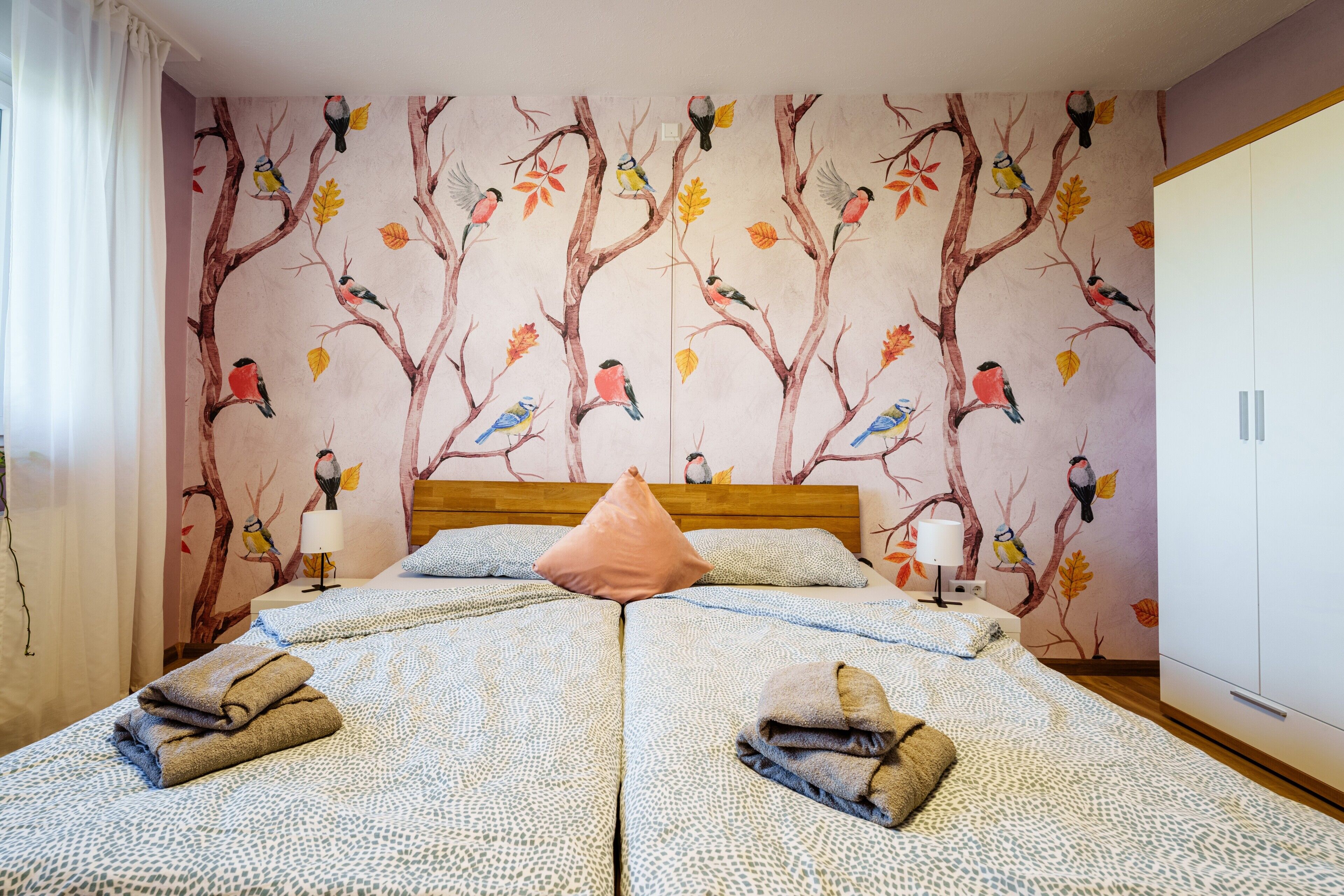 Un lit double avec une couvre-lit à motifs, deux lampes de chevet, une tête de lit en bois et une fresque murale représentant des oiseaux et des feuilles.