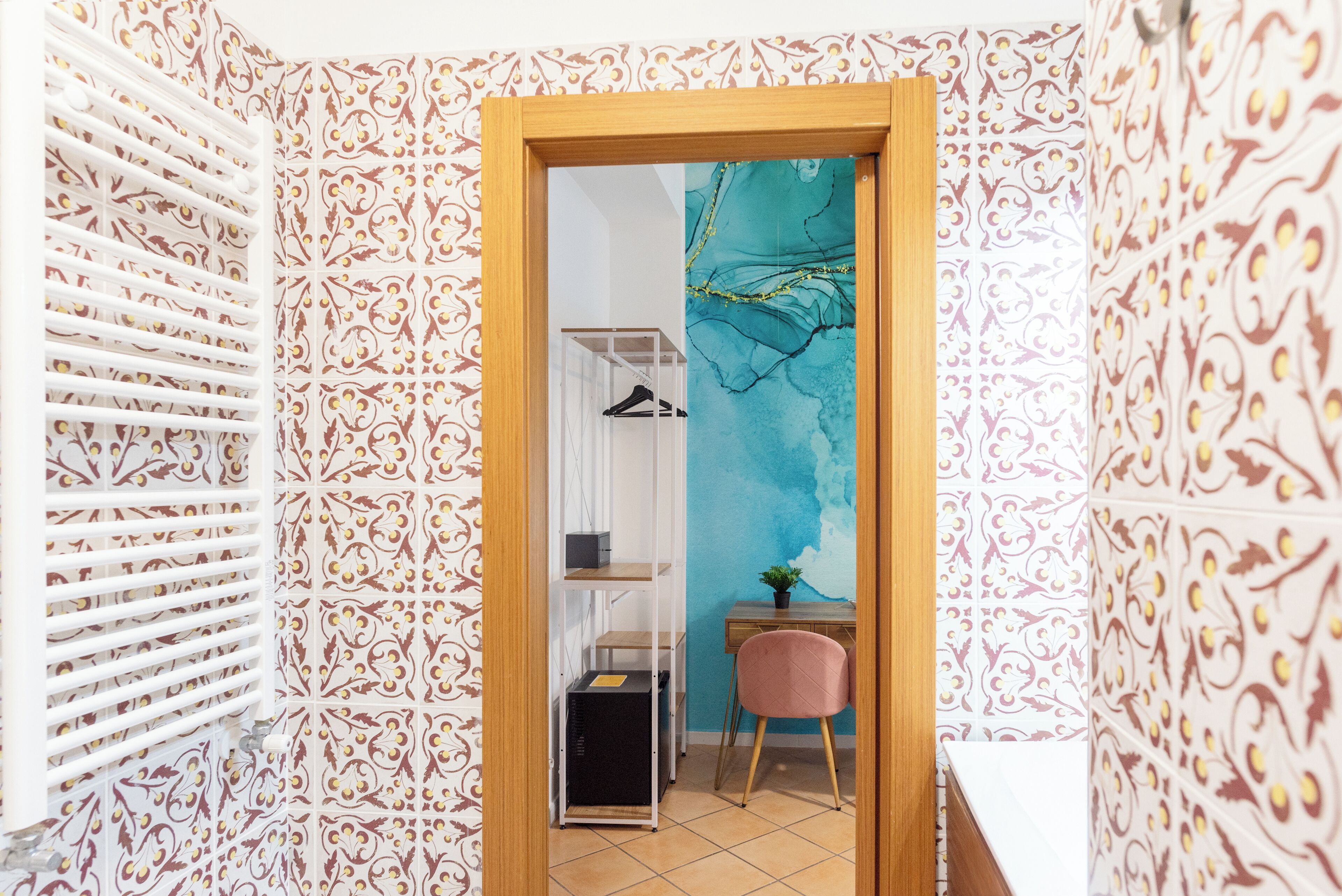 A room with patterned wallpaper, a wooden door, a desk with a chair, and a shelf with a plant.