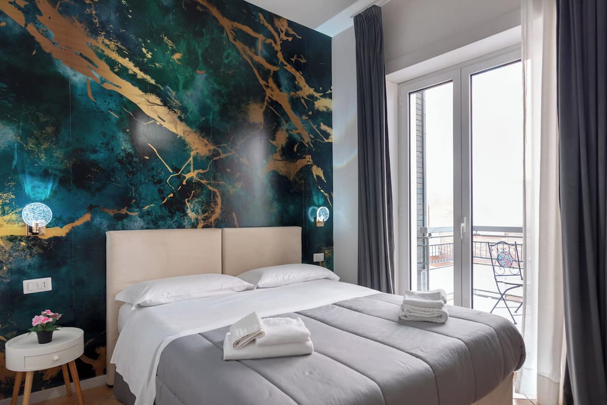 A modern hotel room with a large bed, a round side table, a nightstand, a wall with a unique abstract mural, and a balcony with a view.