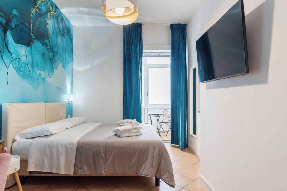 A modern hotel room with a bed, a flat-screen TV, a mural on the wall, and a balcony with blue curtains.