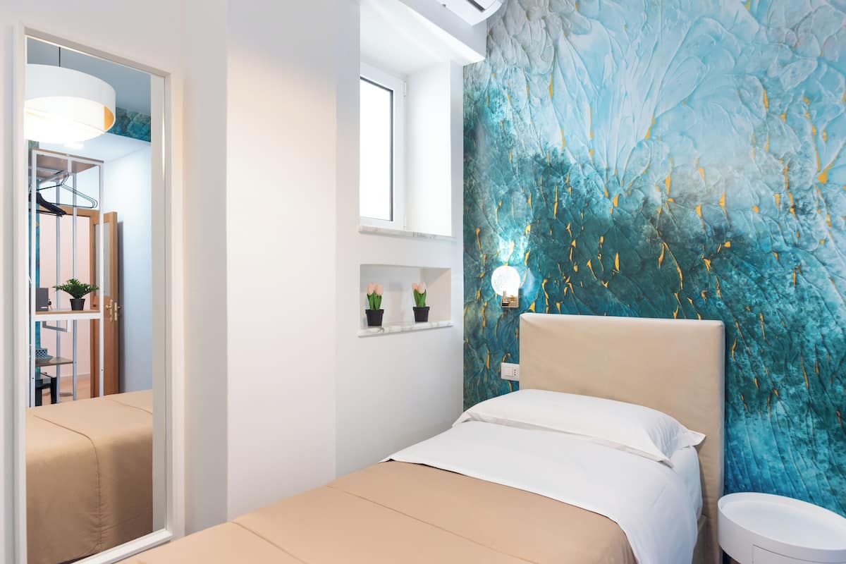 A bedroom with a bed, a nightstand, a wall-mounted lamp, and a window with a view of the outdoors.