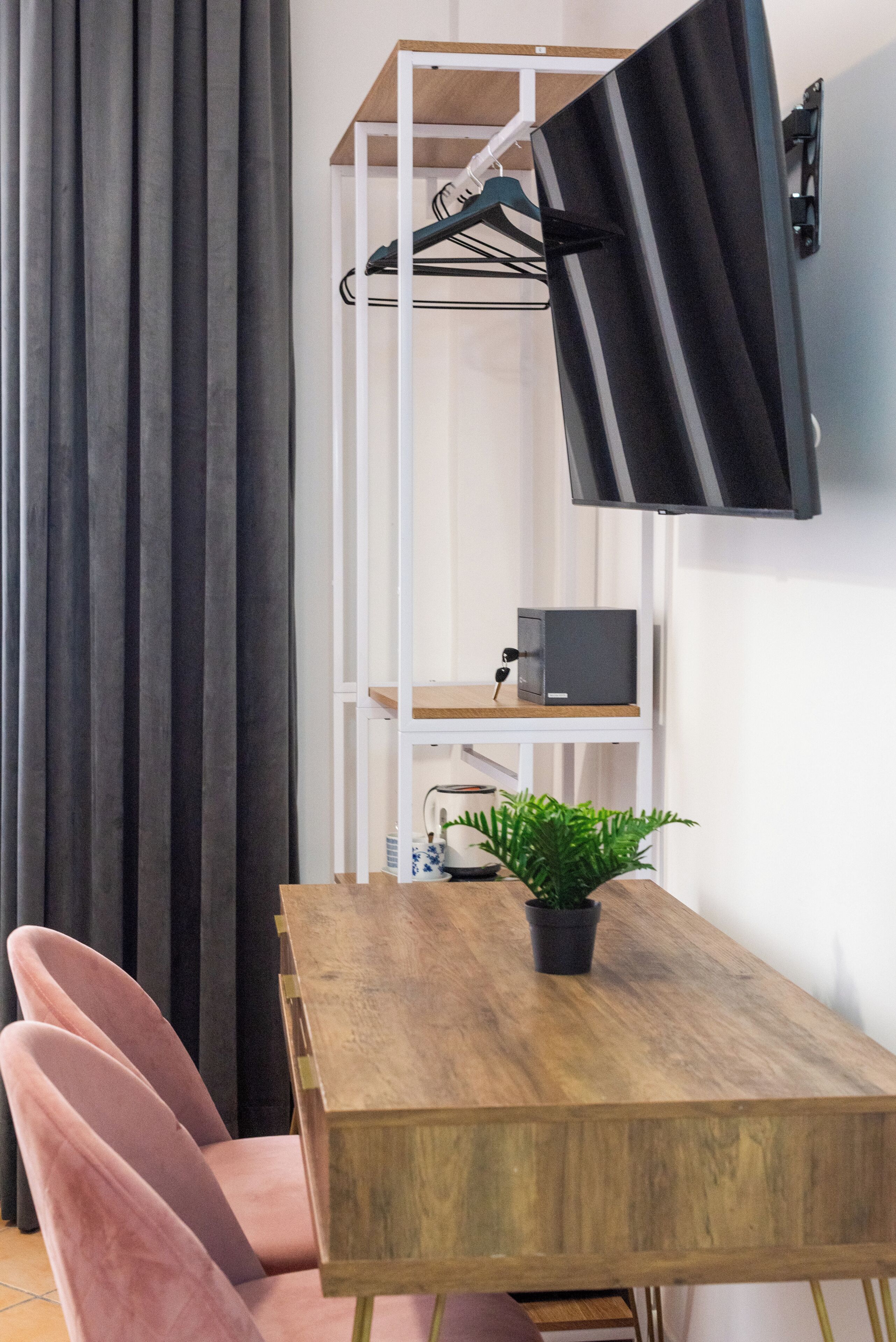 Quarto com mesa de madeira, planta em vaso, cadeira rosa, televisão fixada na parede e um cabideiro.