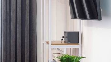 Quarto com mesa de madeira, planta em vaso, cadeira rosa, televisão fixada na parede e um cabideiro.
