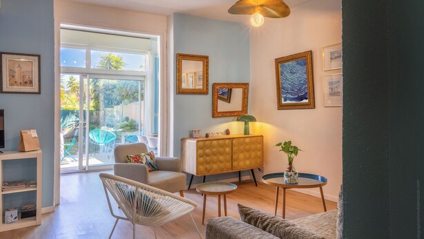 Una sala moderna con sofá, sillón y una mesita con una planta. Se observa un mueble con adornos y un amplio ventanal que proporciona luz natural.