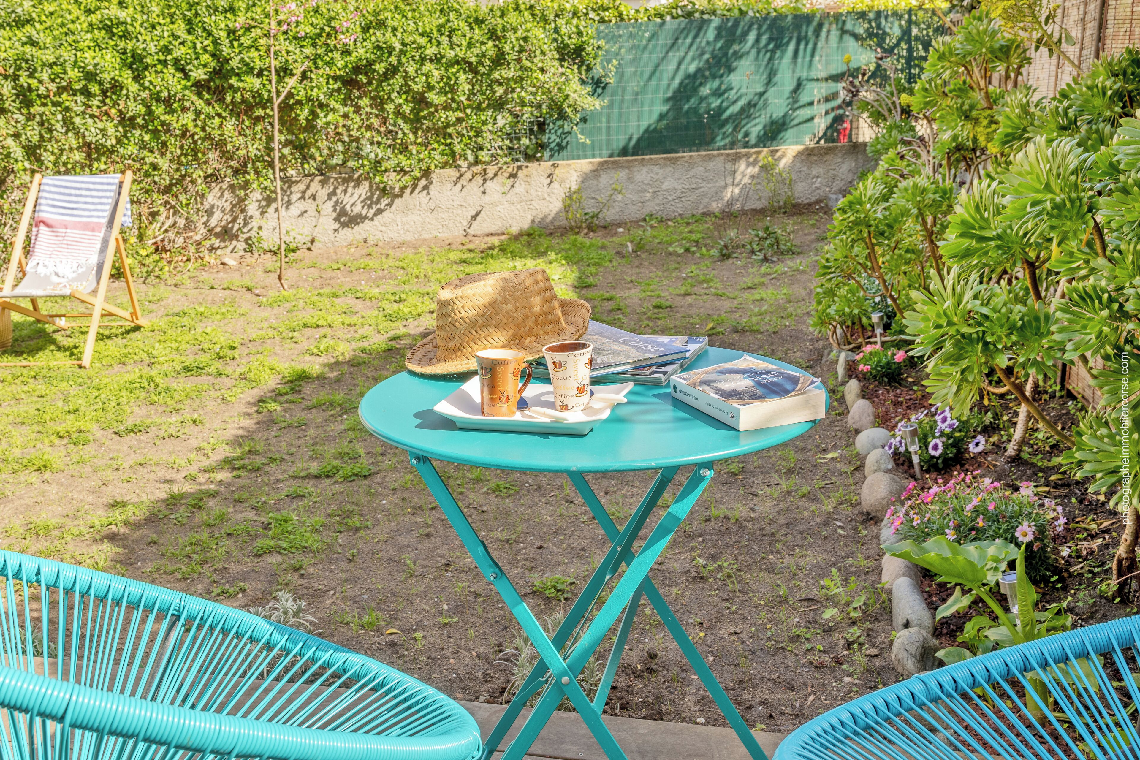 Un pequeño espacio al aire libre con una mesa turquesa, dos sillas, un sombrero de paja, una taza y un libro.