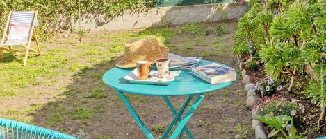 Un pequeño espacio al aire libre con una mesa turquesa, dos sillas, un sombrero de paja, una taza y un libro.
