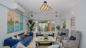 A spacious living room with a blue sofa, patterned armchairs, a glass coffee table, and a large window overlooking a pool. - ELIVAAS Bauhinia 5 BHK Pvt Pool Villa  (Vagator)