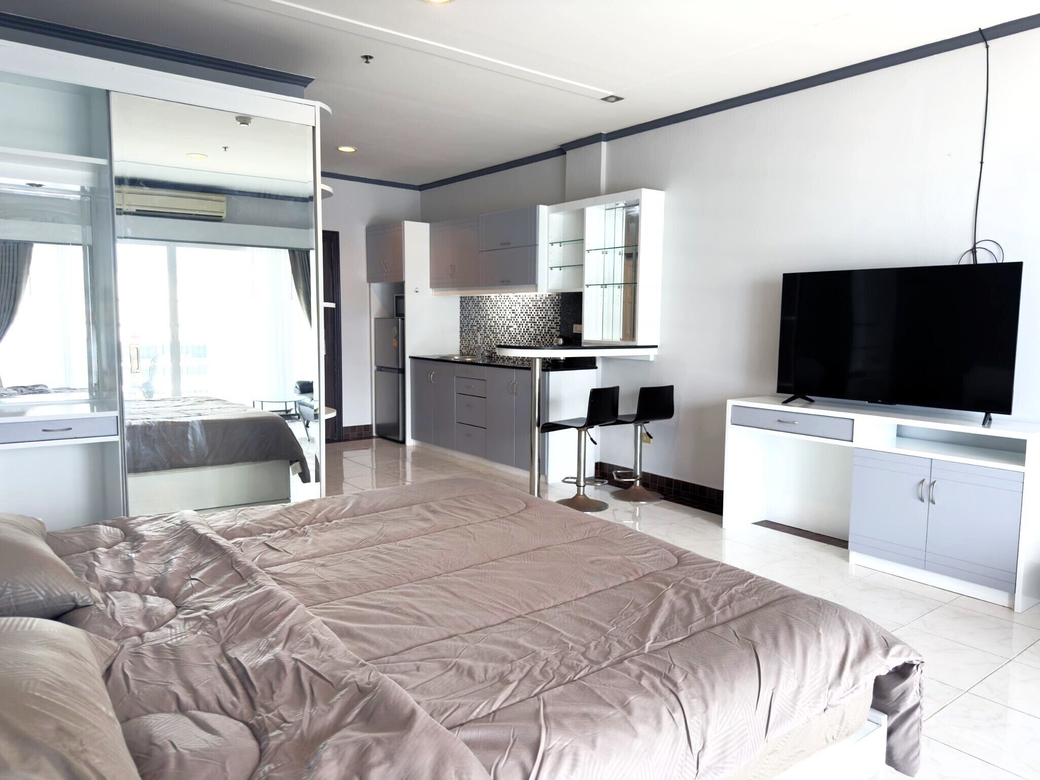A modern hotel room with a large bed, a TV, a kitchenette, and a bathroom with a glass shower.
