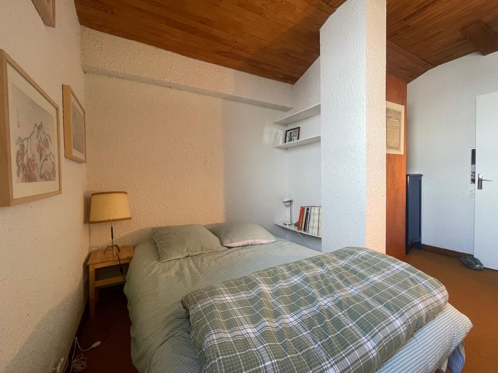 Une chambre à coucher avec un lit, une table de chevet, une lampe, une étagère avec des livres et une porte.