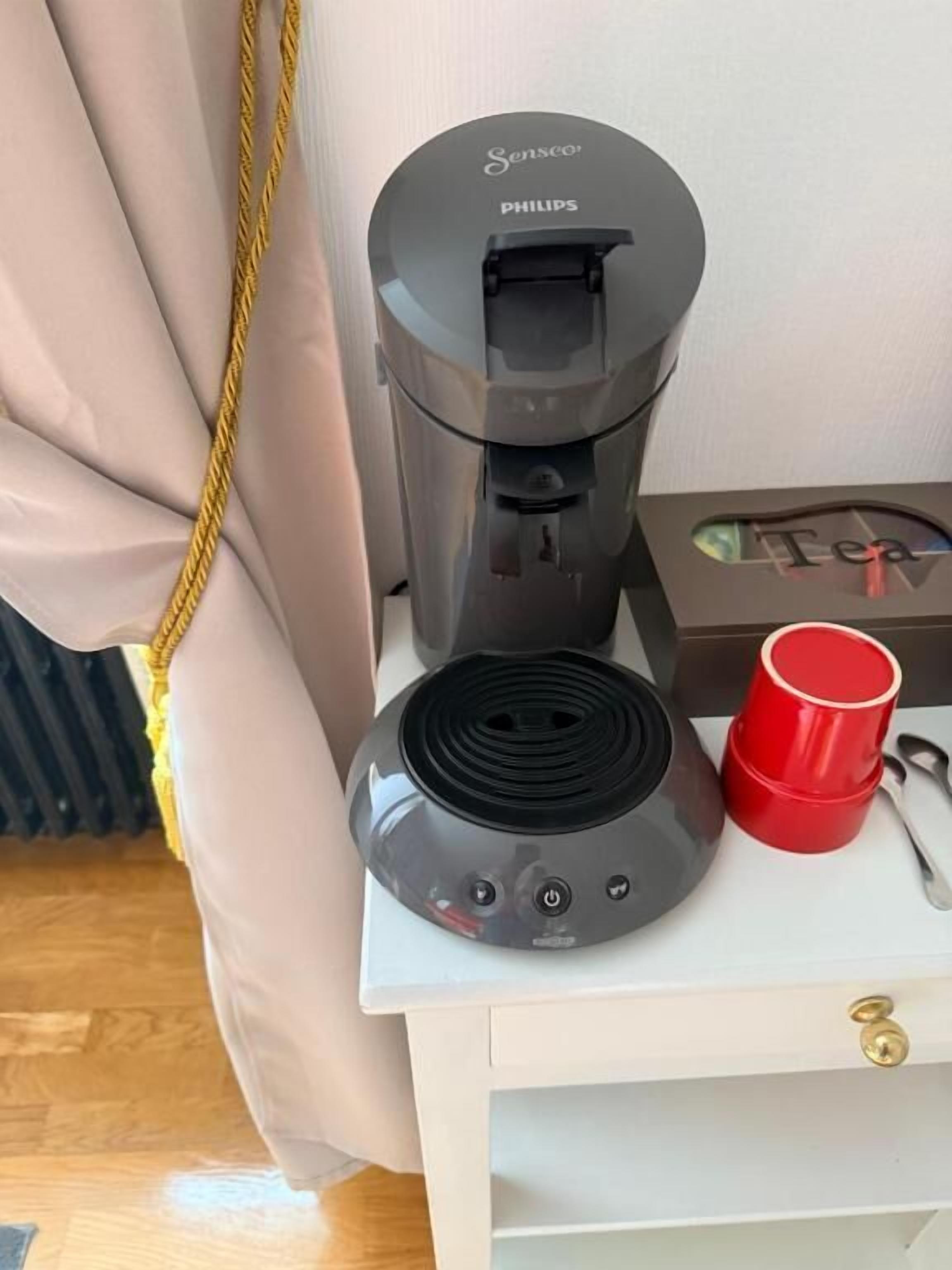 A coffee maker on a bedside table with a red cup and a spoon.