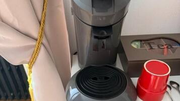 A coffee maker on a bedside table with a red cup and a spoon.