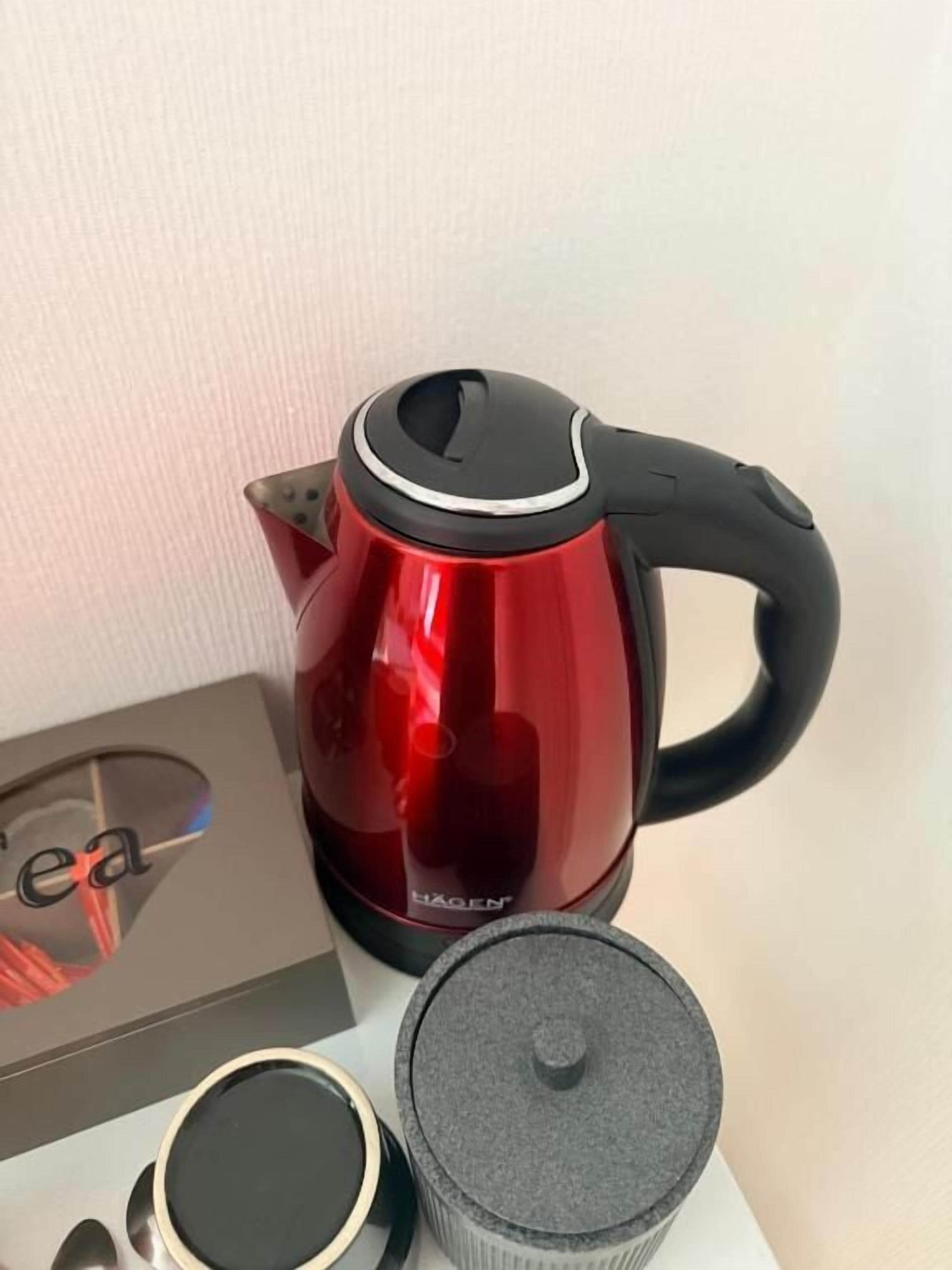 Una tetera roja eléctrica con asa negra, una caja con la palabra “té” y un recipiente gris con tapa.