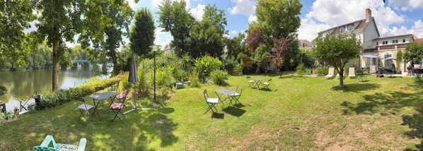 A grassy outdoor area with tables and chairs, a pond, and a house in the background.