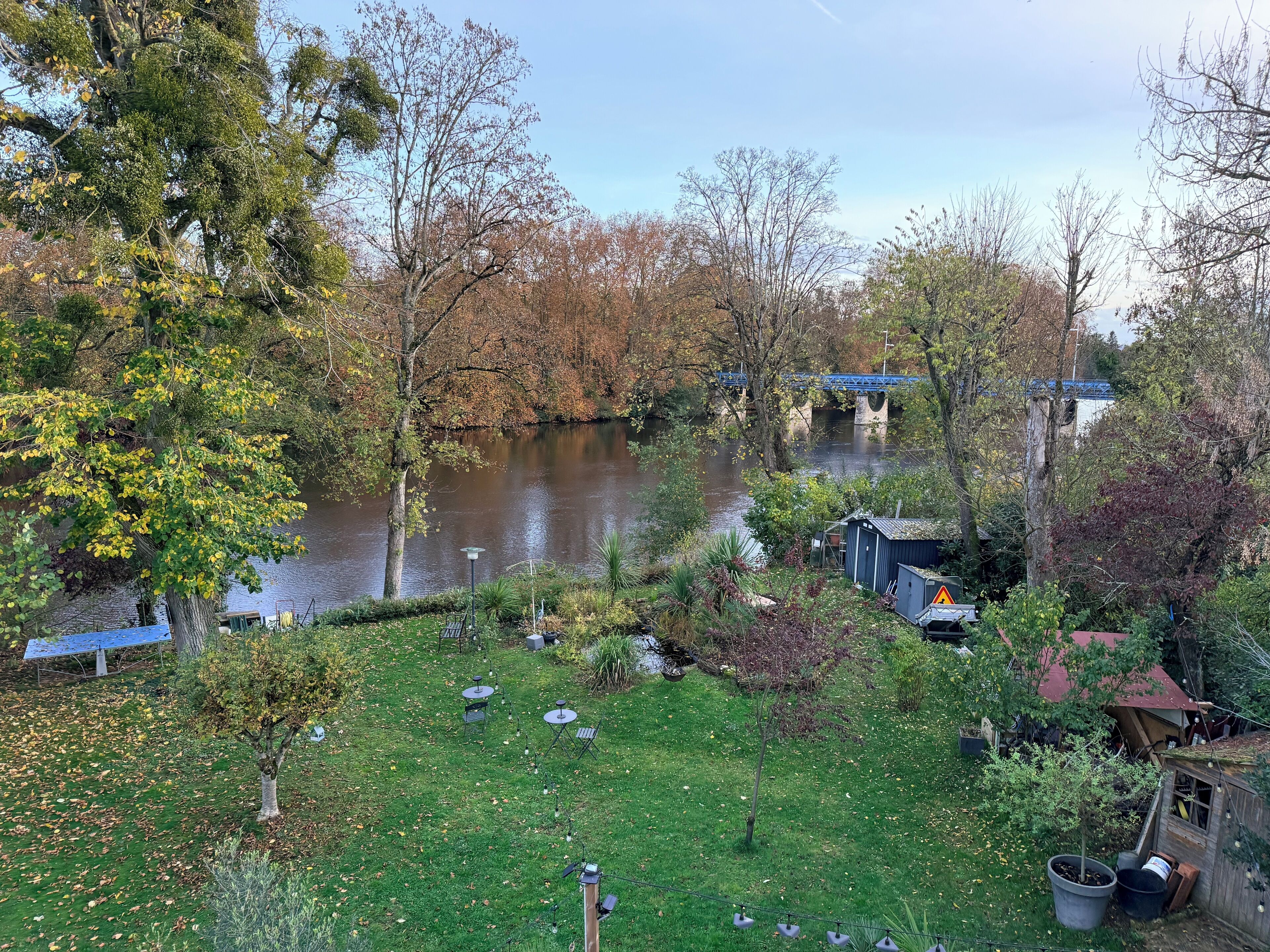 A riverside garden with a small wooden structure, a bridge in the background, and a grassy area with scattered leaves.
