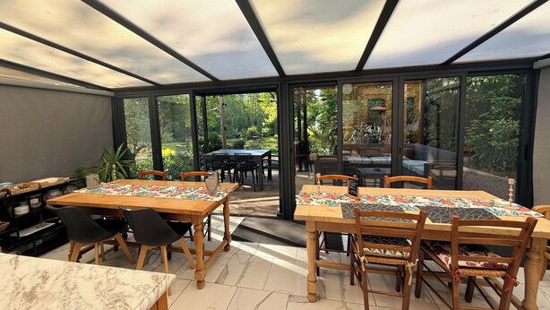 A conservatory with a glass ceiling, wooden tables, and chairs, and a view of the garden outside. - villa du beau port (Cenon-sur-Vienne)