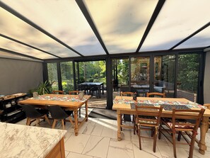 Een conservatorium met een glazen dak, houten tafels en stoelen, en uitzicht op de tuin.