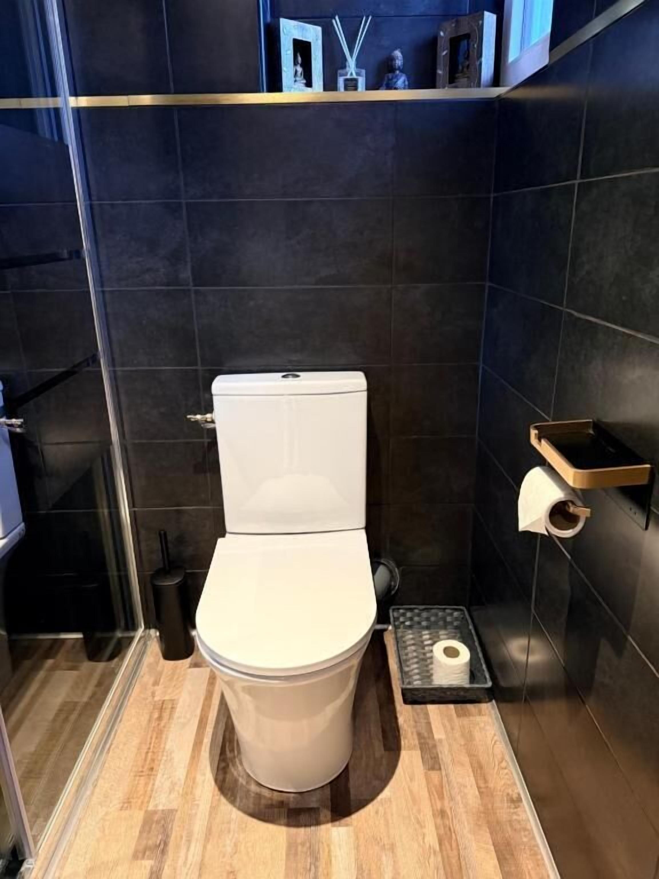 A modern bathroom with a white toilet, dark tiled walls, a wooden floor, and a small shelf with toiletries.