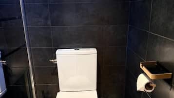 A modern bathroom with a white toilet, dark tiled walls, a wooden floor, and a small shelf with toiletries.