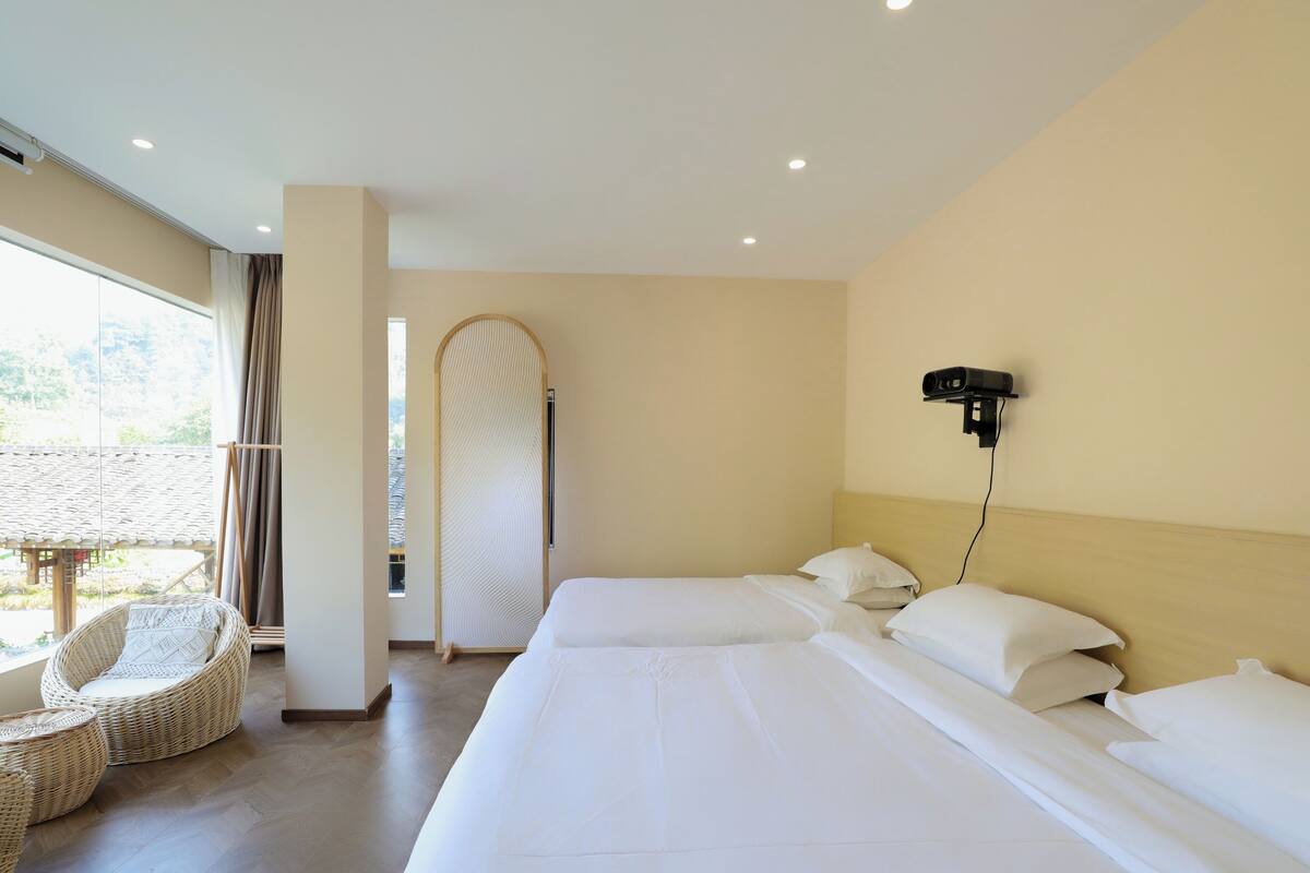 A hotel room with a large bed, a seating area with wicker furniture, a wall-mounted projector, and a view of the outdoors.