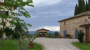 A rural setting with a gravel path leading to a stone building with a tiled roof, surrounded by greenery and a mountain backdrop. - Tenuta Branca – Apartment La Loggia with Pool and Private Terrace in Chianti (San Casciano in Val di Pesa)