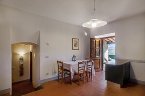 Un espace repas comprenant une table et des chaises, un luminaire suspendu et une vue sur l’extérieur grâce à une porte ouverte.