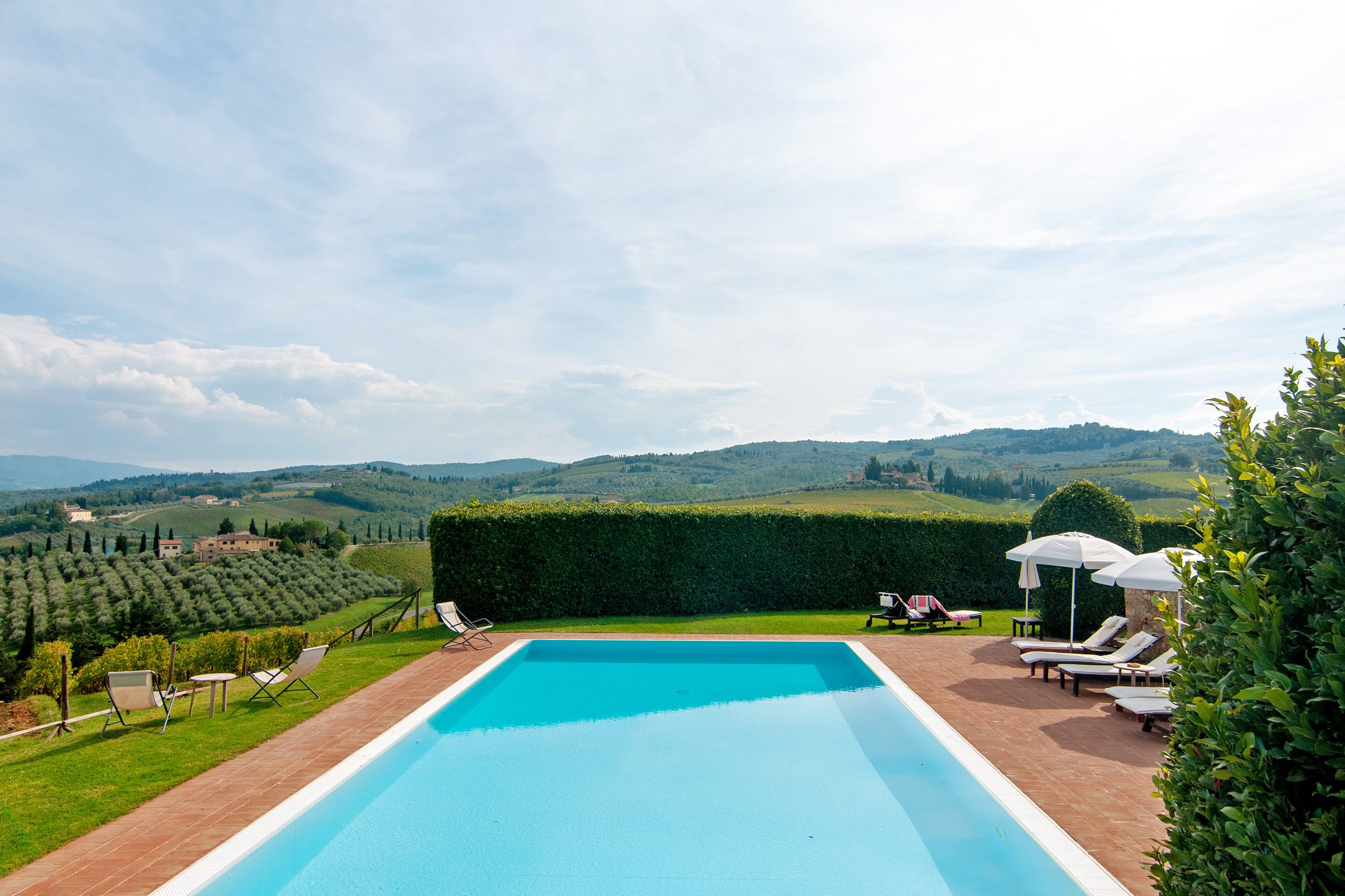 A rectangular swimming pool with a clear blue water surface, surrounded by a brick deck. There are lounge chairs and umbrellas on the deck, and a neatly trimmed hedge to the right. In the background, there are rolling hills and a clear sky.